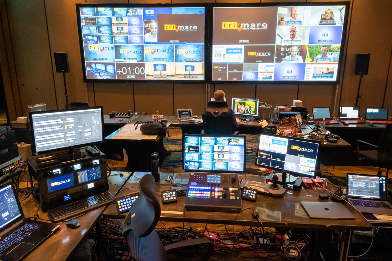 The view behind the scenes at a virtually broadcasted Tri-Marq event. There is a large projector screen in the front of the room with Zoom cameras, timers, and other broadcasted materials.