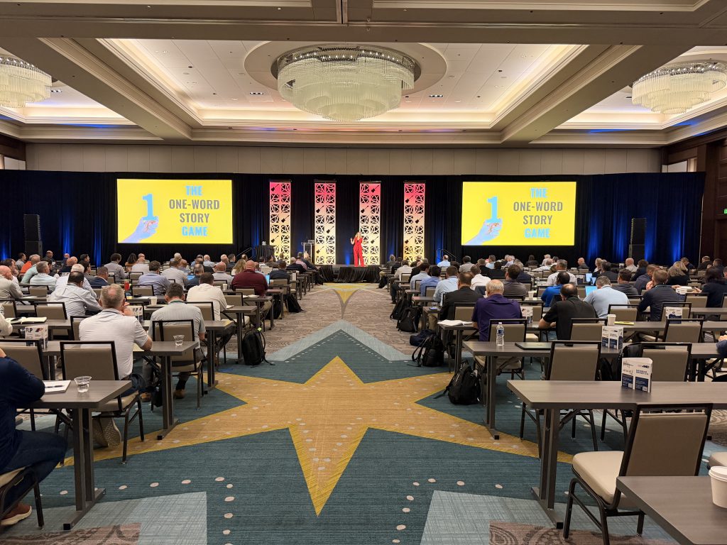 For gallery page: A view from back of house during the General session. Features a presenter speaking with content on both projection screens.