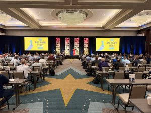 A view from back of house during the General session. Features a presenter speaking with content on both projection screens.
