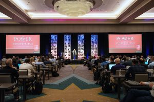 A view from back of house as a presenter is speaking. Features speaker in center-stage with content on both projection screens.