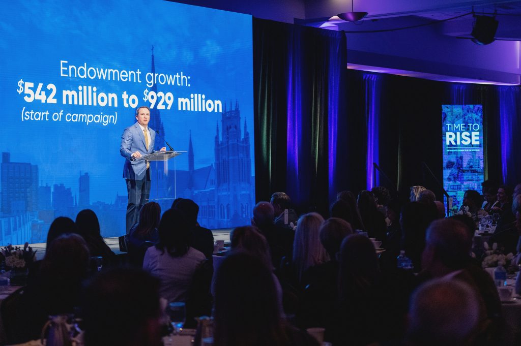 A view of a speaker presenting on stage in front of an LED board with statistics describing money raised for scholarships. 