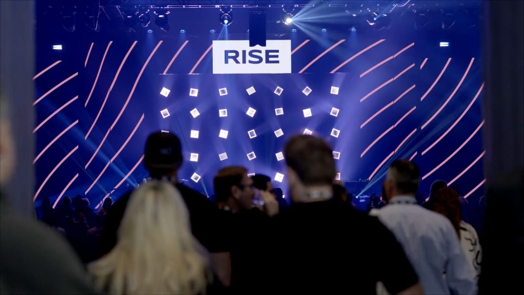 For gallery page: A thumbnail from the Real Brokerage RISE Behind-the-scenes case study video. Features a crowd of people walking into the general session room with the stage setup in the background. 