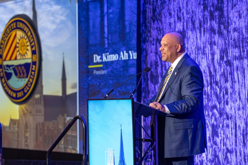 For gallery page: A photo from the Marquette University Scholarship Event. Features Dr. KImo Ah Yun speaking on stage behind an LED podium. 