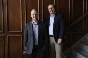 Tri-Marq managing partners, Keith and Eric, pose in business professional clothes in front of a wooden wall.